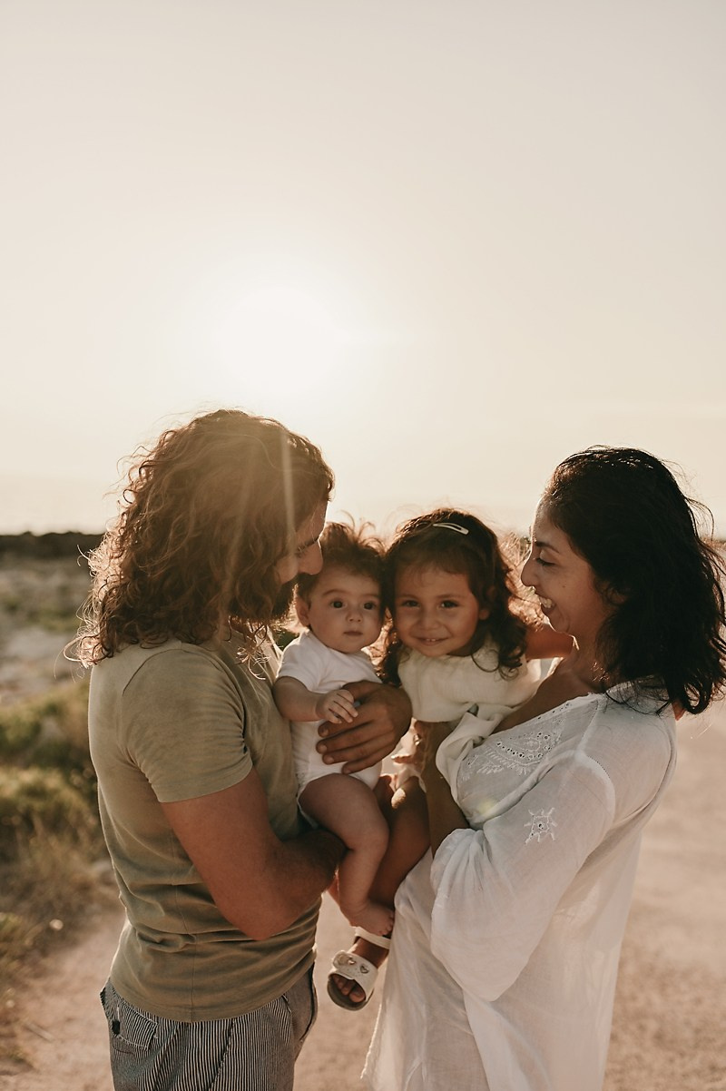 A mother and father smiling as their two daughters cuddle and touch their baby sibling at sunset.