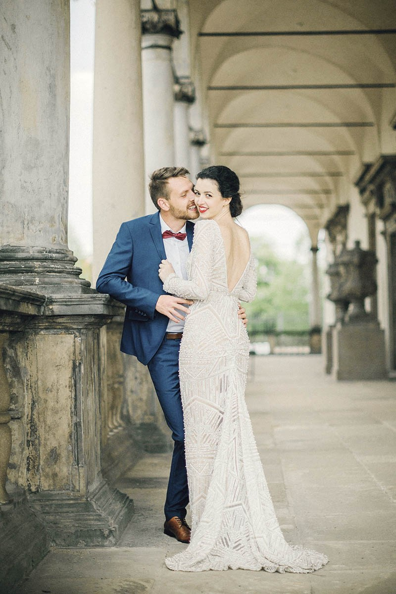 A man lovingly embraces his partner as she looks over her shoulder towards the camera's gaze at the Belvedere Palace in Prague.
