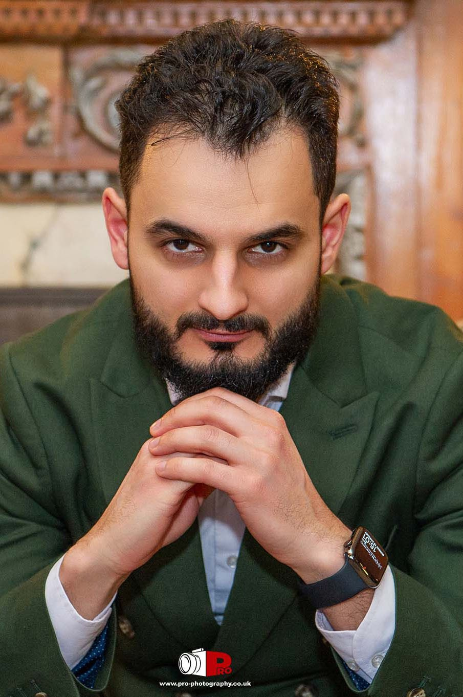 A stylish man in a green suit posing confidently with hands clasped in front of his face, set against a classic wooden fireplace background.