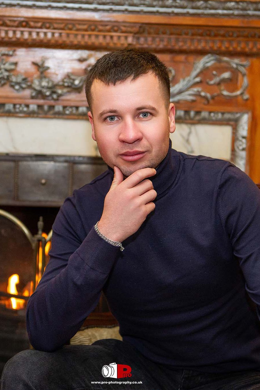 A man in a navy sweater posing by a warm fireplace, showcasing relaxed and inviting portrait photography.