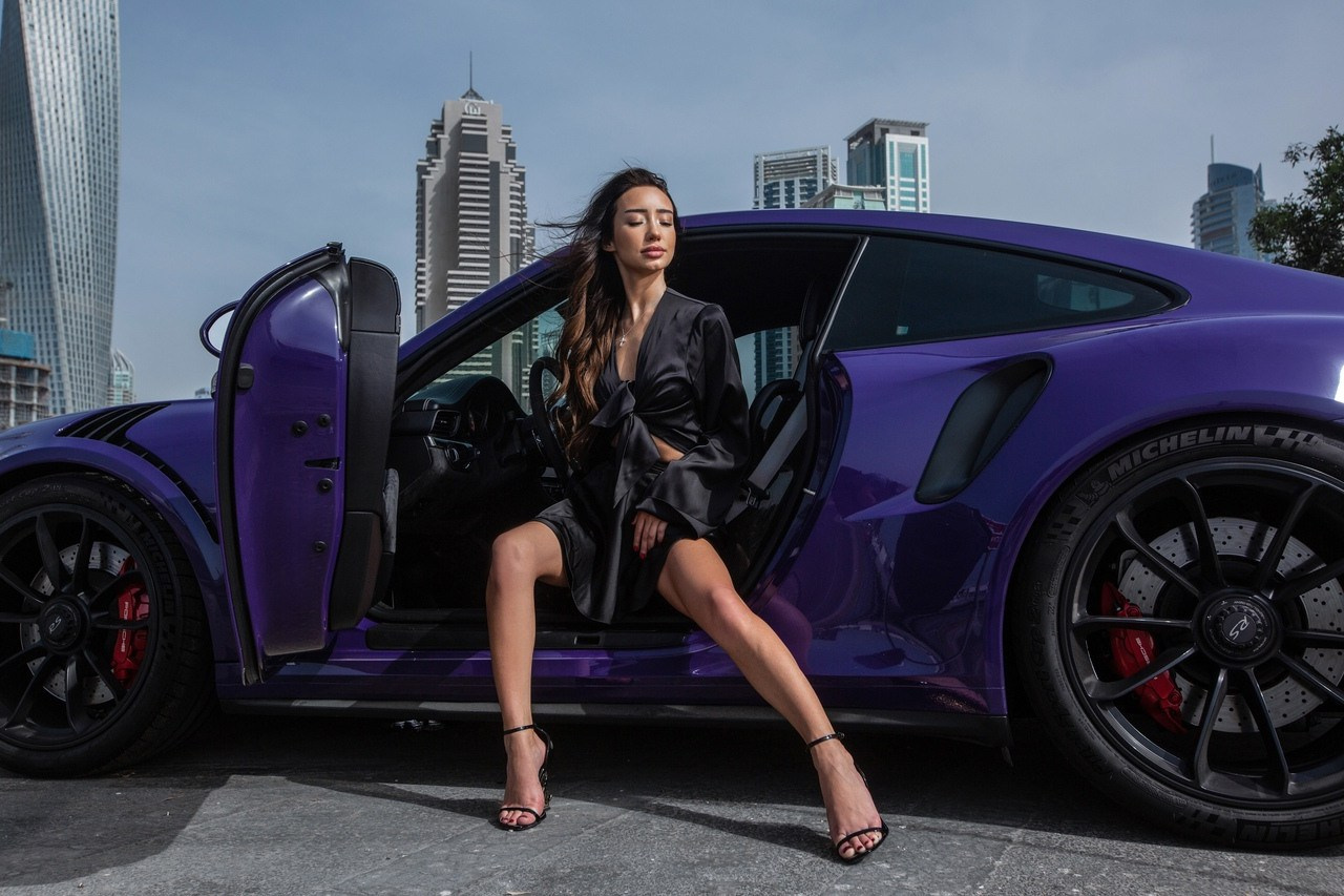 A model in elegant black attire sits inside a blue car with Dubai’s skyscrapers in the background