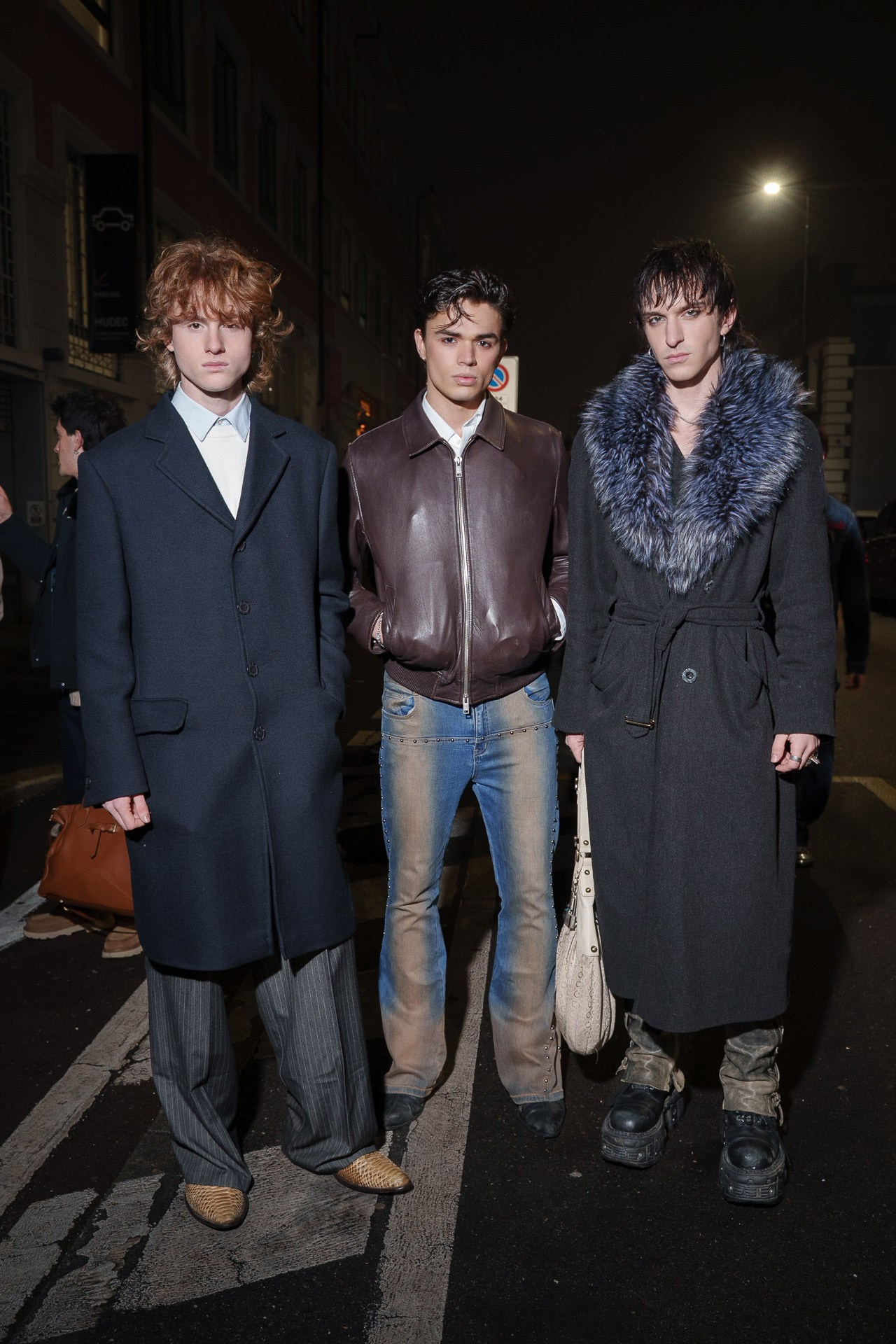 Three male fashion models wearing winter coats and leather jacket photographed at night on a city street during fashion week