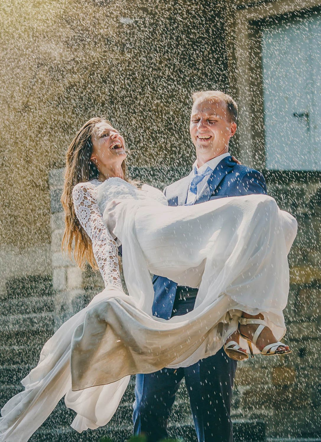 A groom carries his bride in his arms as they are engulfed by a water sprinkler during their hot summer wedding day.