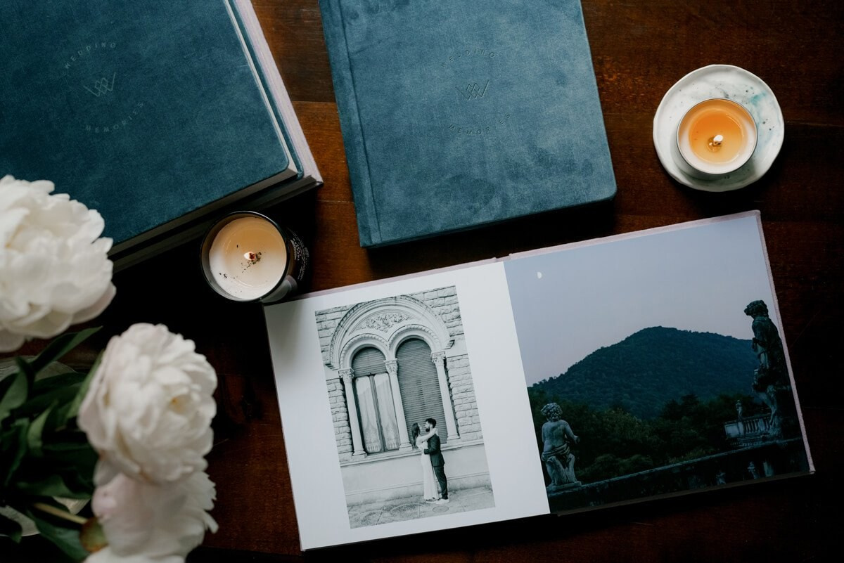 Luxury fine art wedding album open to black-and-white destination wedding portraits, styled alongside dark velvet album covers, white peonies, and lit candles
