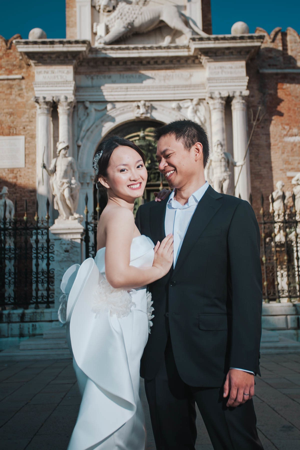 Thai bride smiling as she embraces her partner in front of a historic gate in Venice's Arsenal.