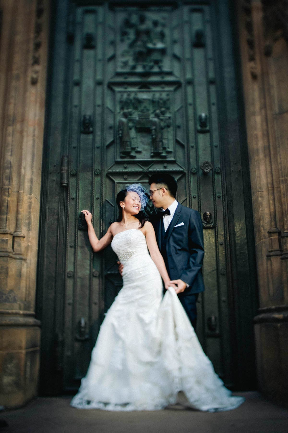 The NYC bride Jen wearing a birdcage veil and a vintage dress is embraced by her smiling, tuxedo-wearing groom Dennis in front of the historic bronze doors of St. Vitus Cathedral.