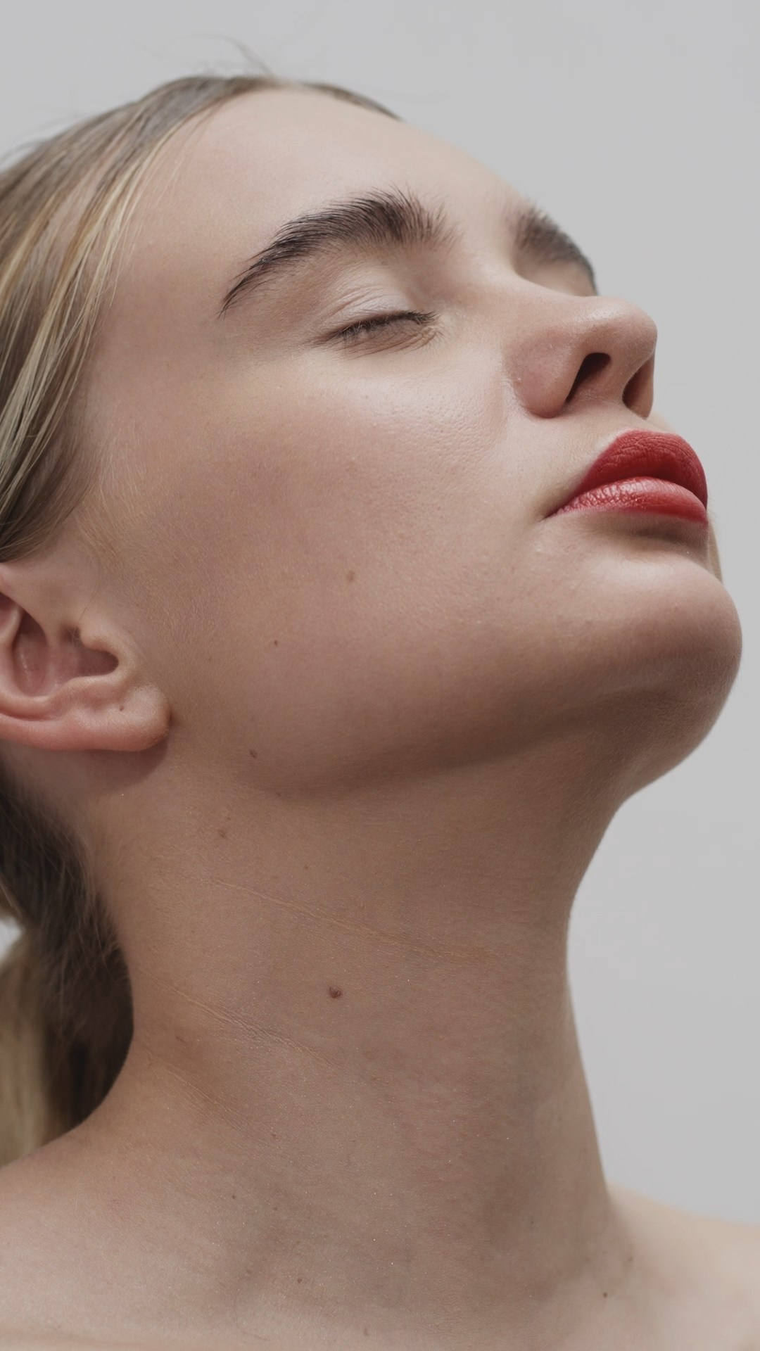 A close-up side profile of a woman with her eyes closed, showcasing her smooth skin and bold red lipstick, exuding a serene and elegant mood.