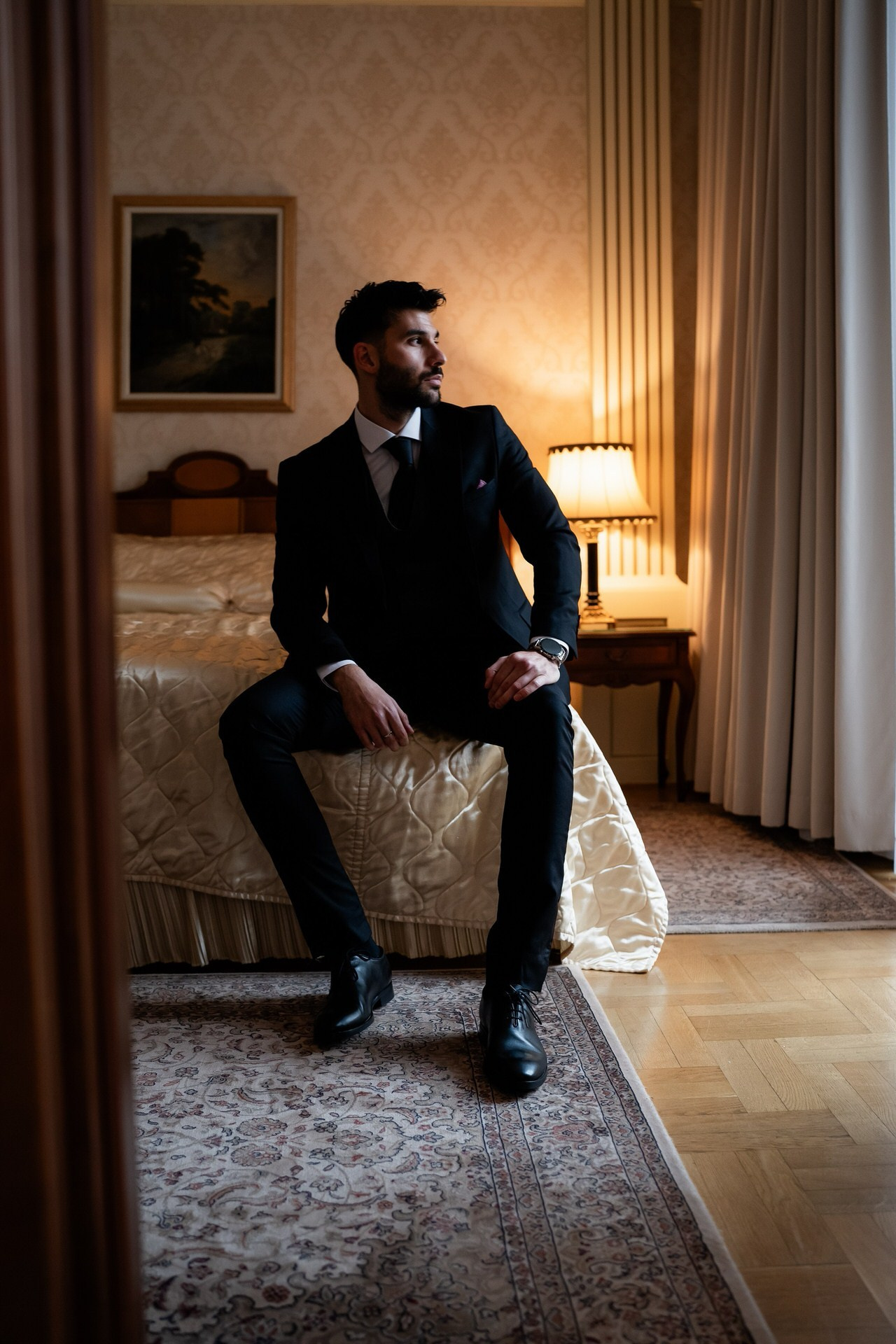 Mladoženja u tamnom odelu sedi na ivici kreveta u elegantnoj hotelskoj sobi, gledajući ka prozoru u toplom ambijentalnom svetlu.