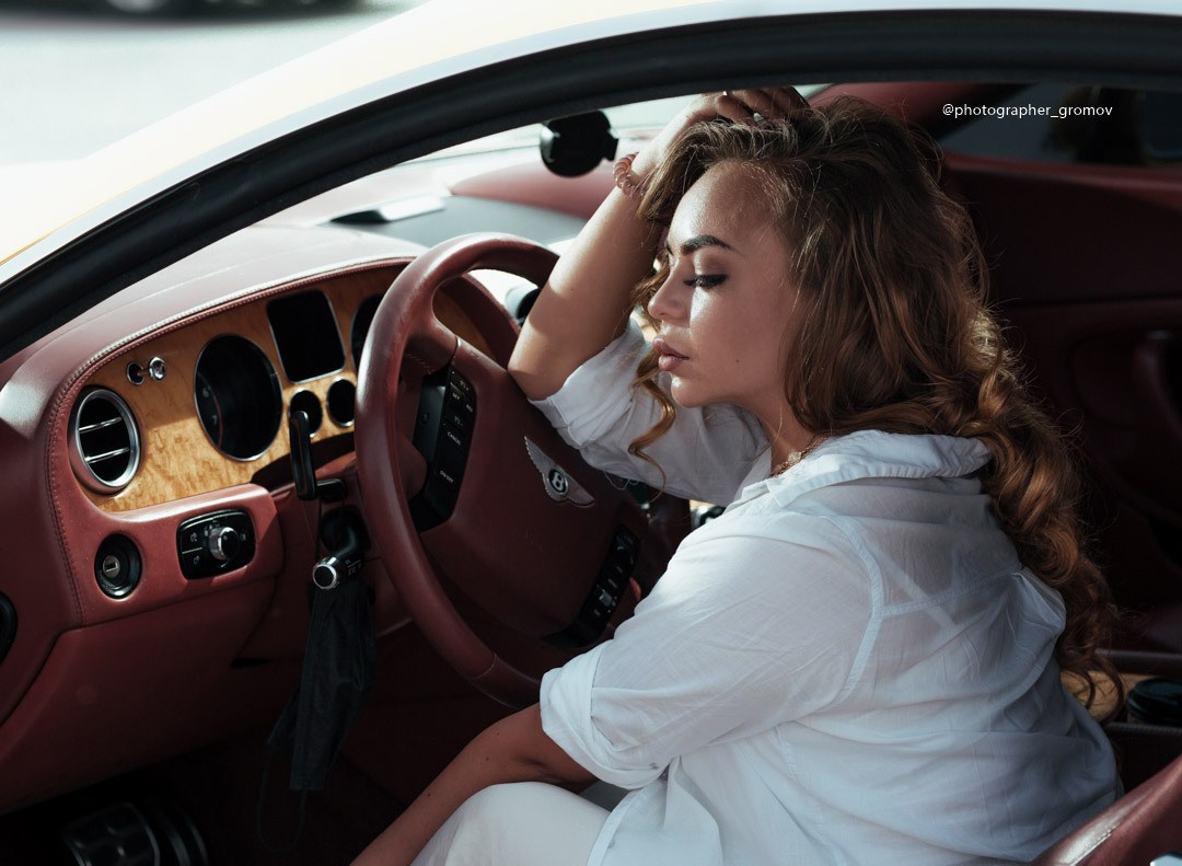 A very tired girl rests her hand on the steering wheel inside the burgundy-colored interior of an exclusive Bentley