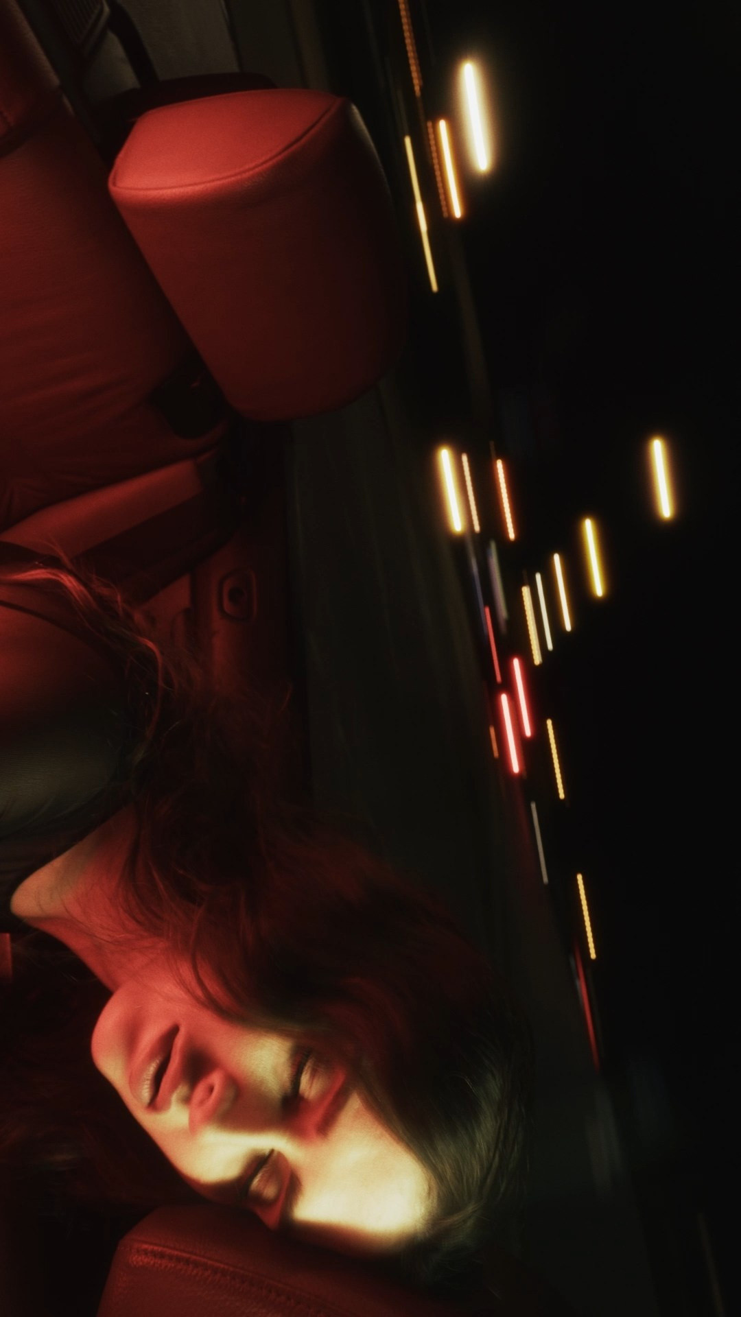 A person reclining inside a car at night, illuminated by warm red light. Their eyes are closed, and their hair flows loosely, with blurred streaks of city lights visible through the car window in the background, creating a dreamy and cinematic feel.
