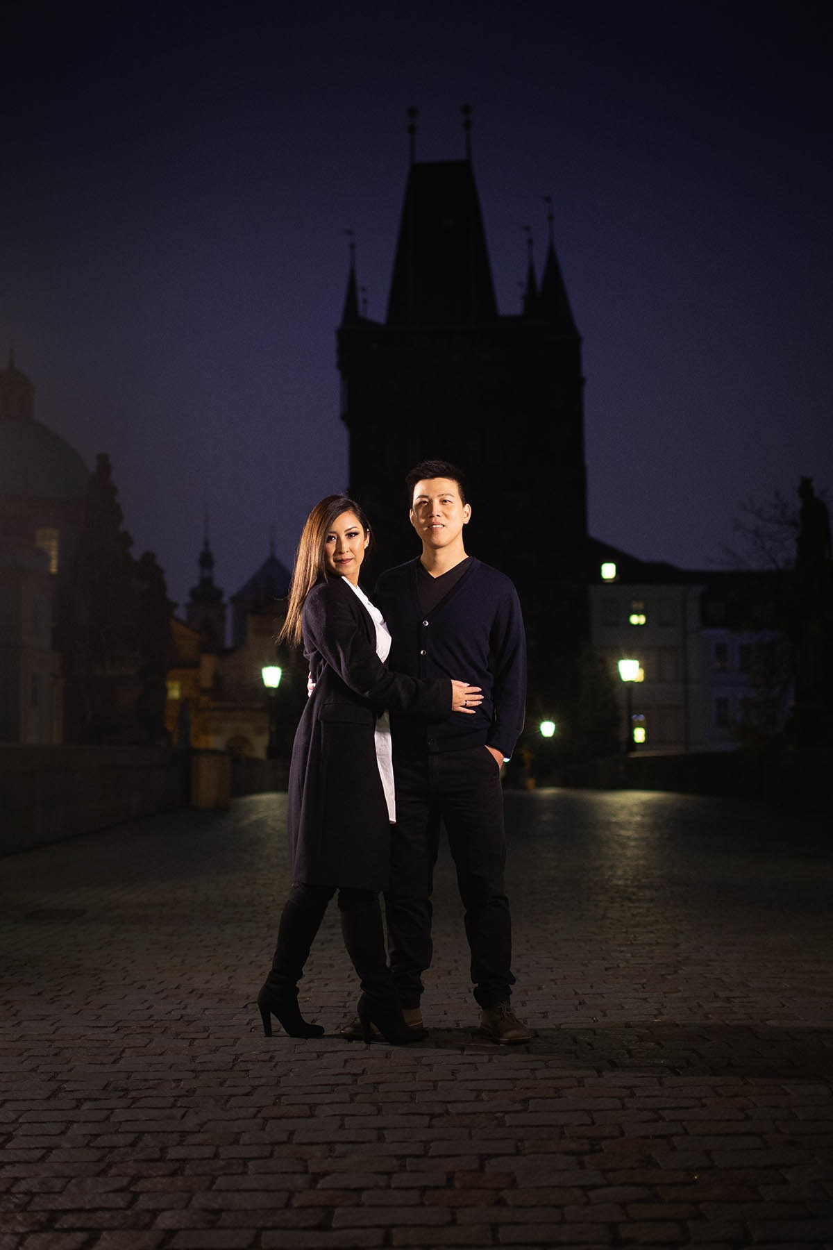 A man and woman stand atop the Charles Bridge at night as the gothic tower is nearly silhouetted against the nigth sky