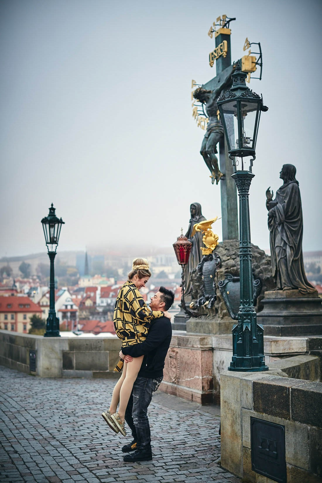 Man lifts woman Calvary statue misty Charles Bridge dawn