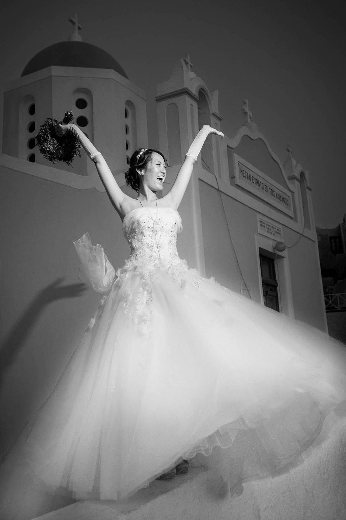 A radiant Japanese bride throws her arms up in celebration while holding her bouquet against the blue sky overlooking a church in Santorini.