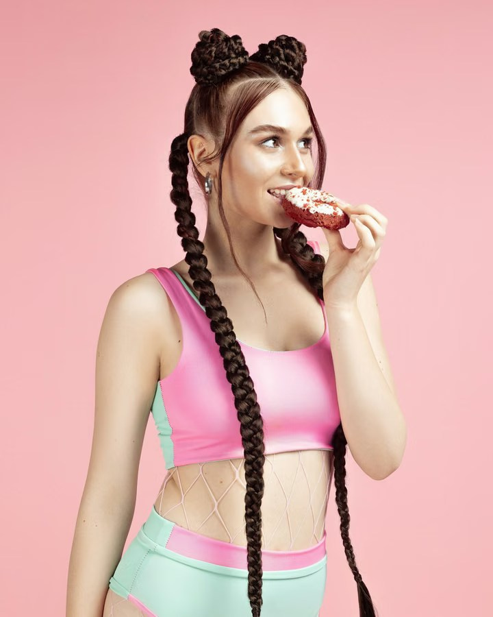 Woman with long braids in fitness wear eating a doughnut against a bright pink backdrop