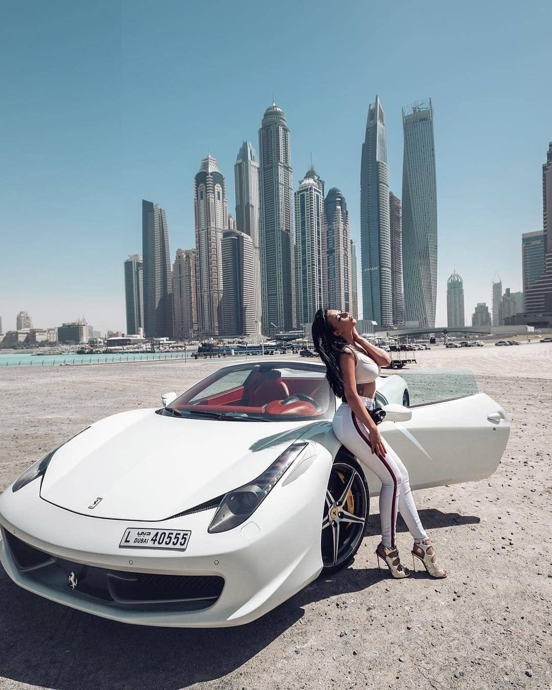 A beautiful model sits on a white Ferrari car. With the hood open against the backdrop of Dubai skyscrapers during a beautiful sunset