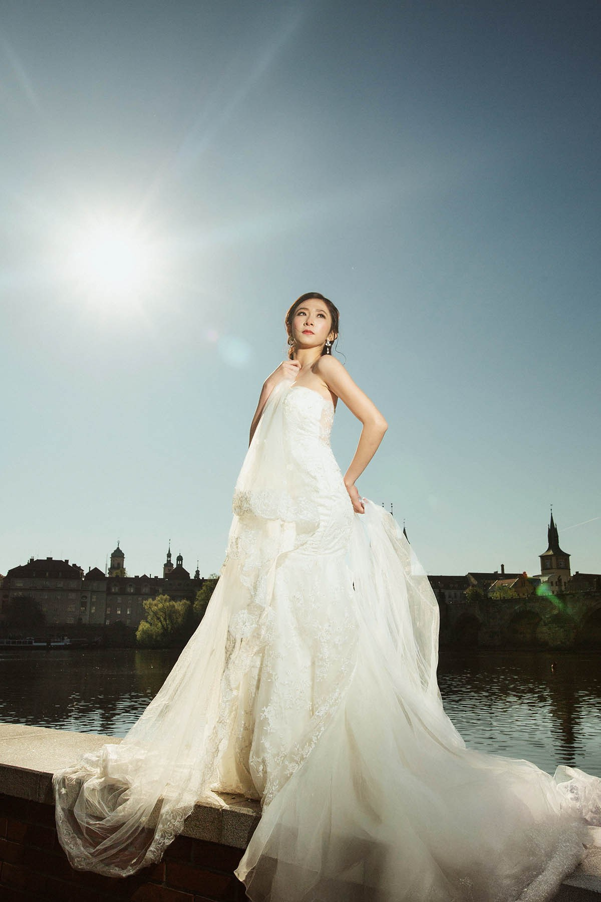 Bride with a sunflare behind her, posing in her wedding dress along the riverside in Prague.