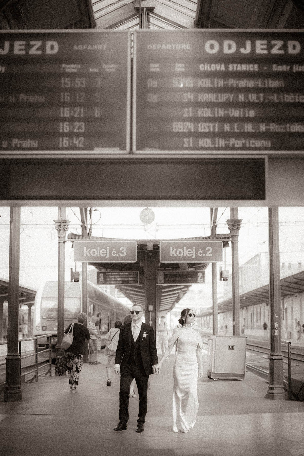 Stylish newlyweds pausing beneath train departure boards at Praha-Masarykovo Station, Prague