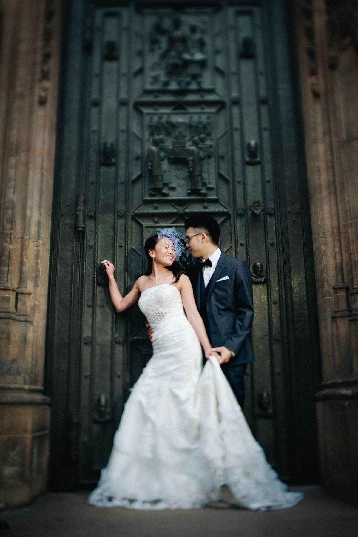 Radiant Asian woman wearing a wedding dress and a bird-cage veil smiles as she is lovingly embraced by her tuxedo-clad groom in front of the historic bronze doors of Prague's St. Vitus Cathedral