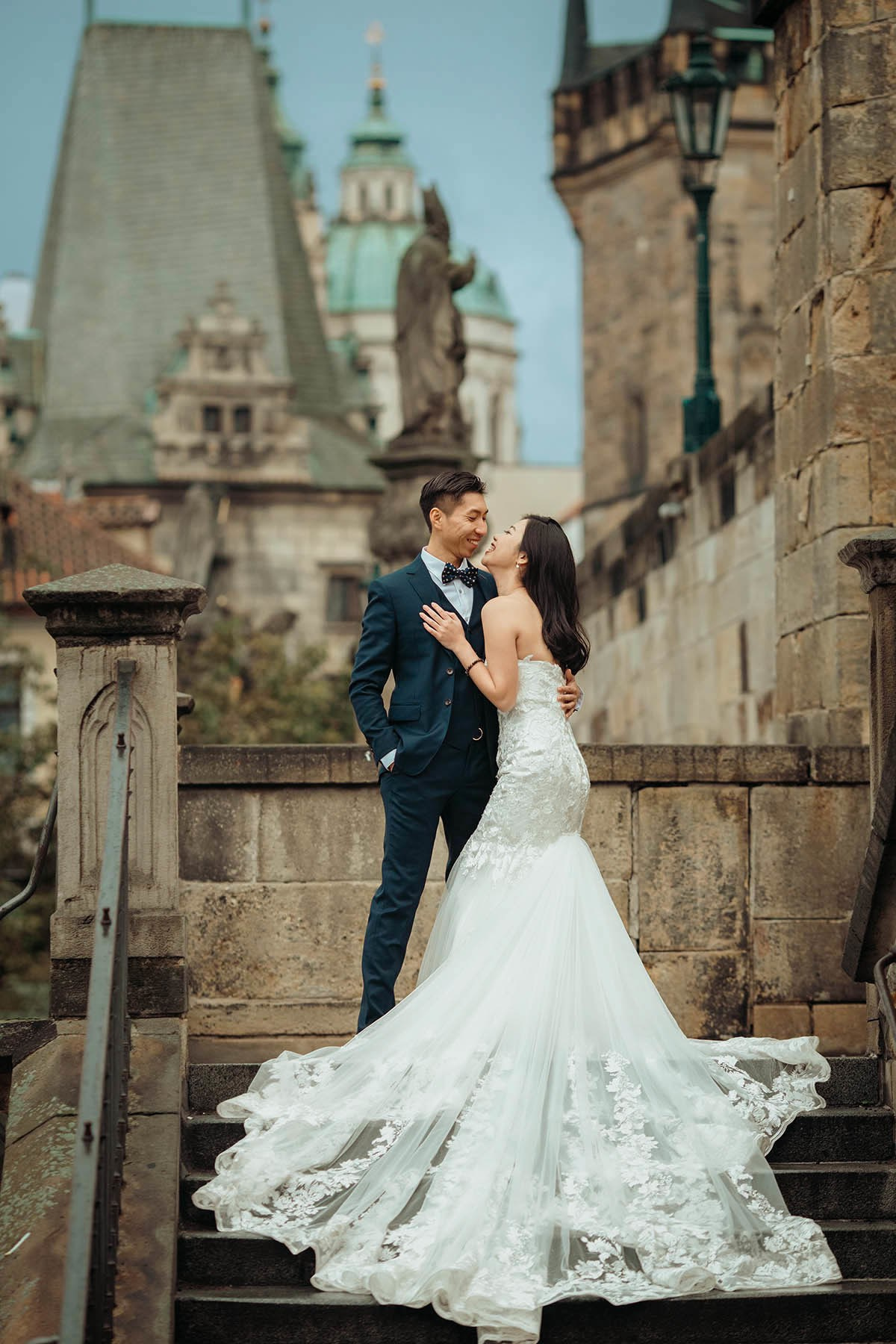 As the gothic towers of the Charles Bridge loom above, a smiing Asian bride is embraced by her tuxedo-wearing groom atop the Kampa steps in Prague
