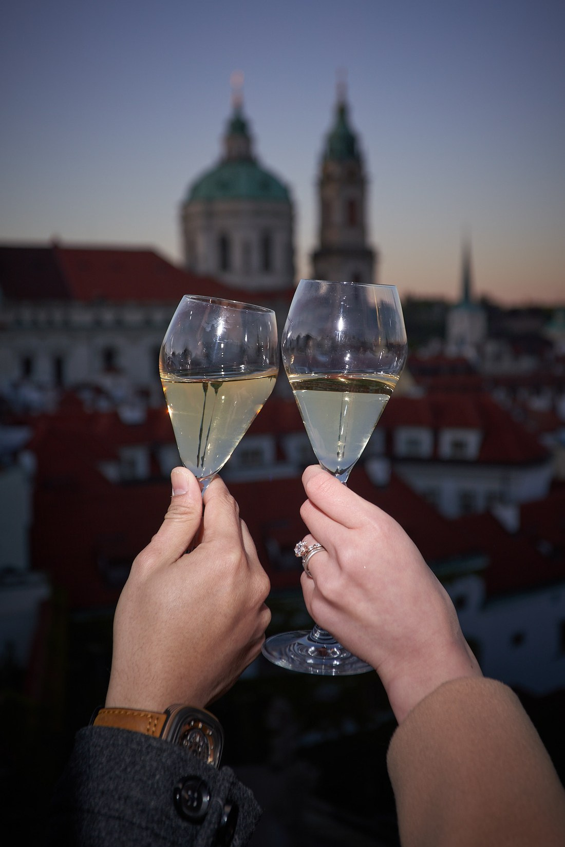 Two hands holding champagnge flutes against the spires of St. Nicholas as the engagement ring reflects the light from the flash.