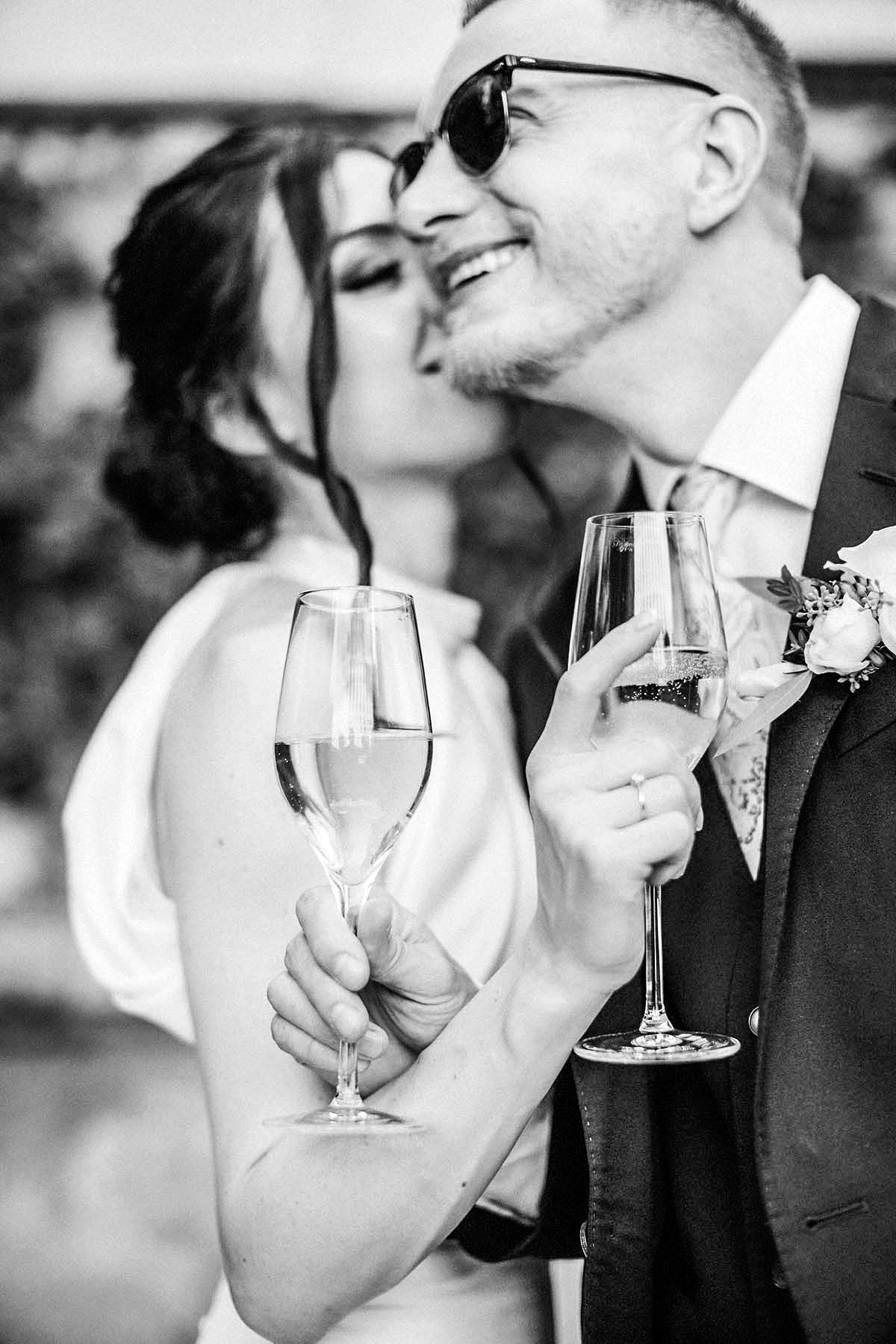 Joyful dark-haired bride kisses the cheek of her rayban sunglass wearing blonde groom while holding their champagne flutes in Prague.