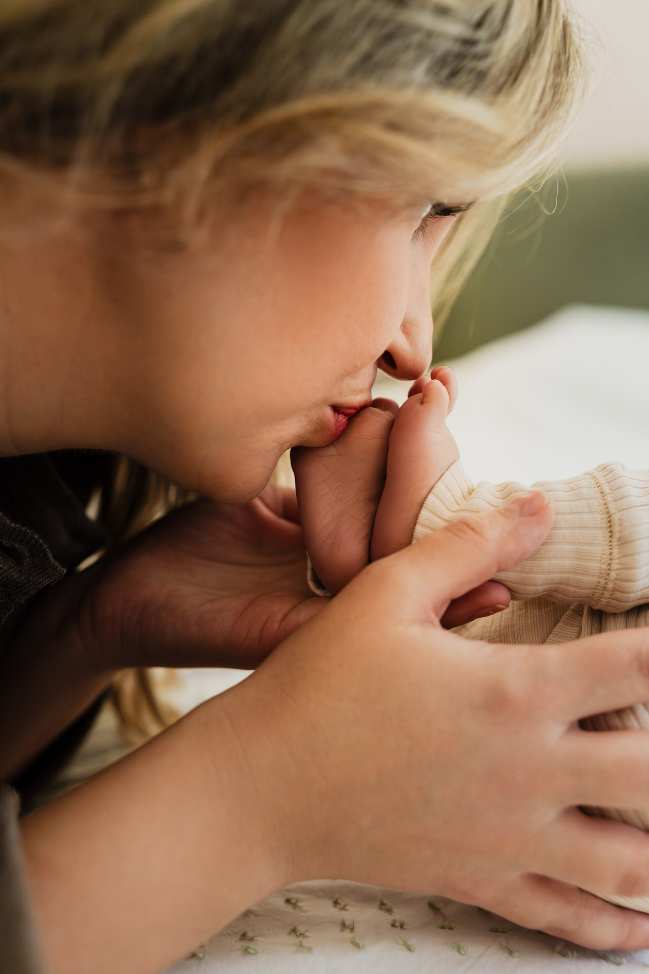 Mama nežno ljubi stopala svoje bebe koja leži na mekanom pokrivaču.