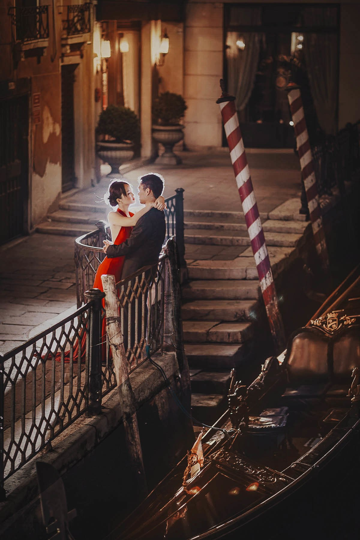 Thai woman in skin-tight red dress lovingly embraced by partner near gondola at night Venice.