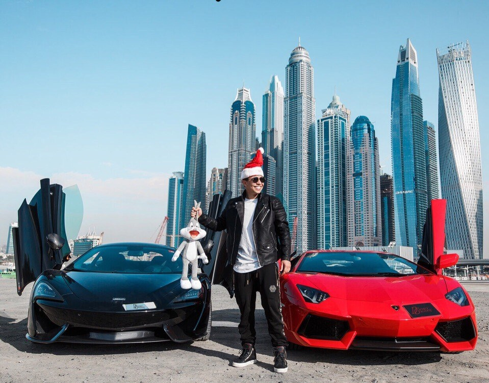 A man stands next to a black McLaren and a red Lamborghini, wearing a Santa hat, with Dubai’s skyscrapers in the background