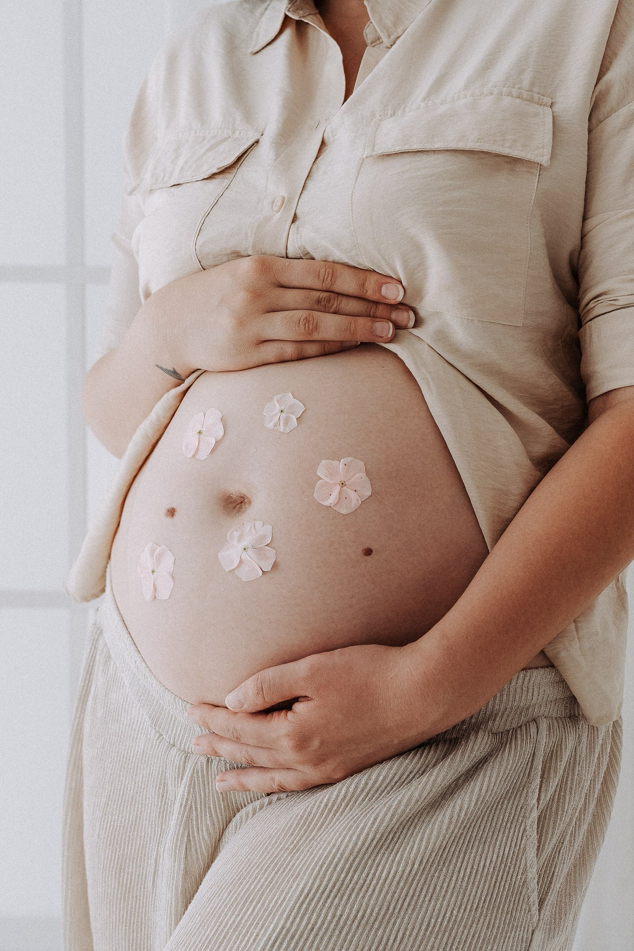 Krupan kadar trudničkog stomaka koji žena нежно drži rukama, sa nekoliko svetloružičastih latica položenih preko kože u svetlom enterijeru.
