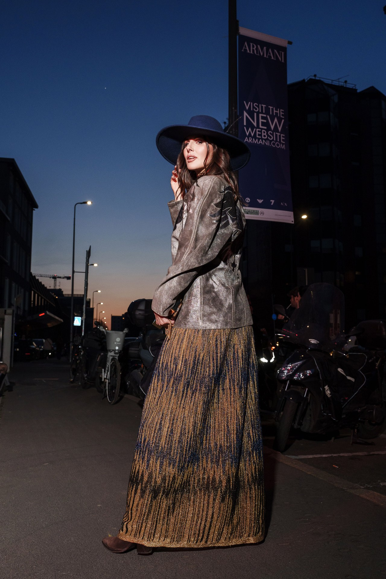 Elegant evening street style portrait of a woman in a wide-brimmed hat and a shimmering gold and blue long skirt at Milan Fashion Week.