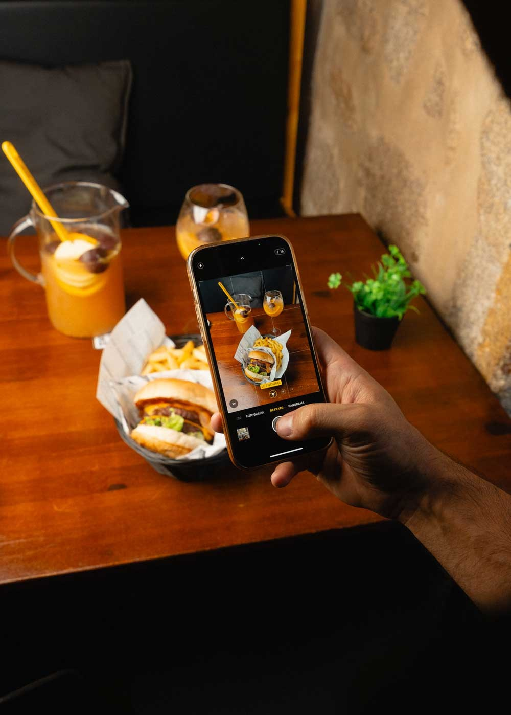 Fotografia de lifestyle e redes sociais de uma mão a usar um smartphone para fotografar um hambúrguer e uma bebida numa mesa de madeira, produzida pela Khave Media