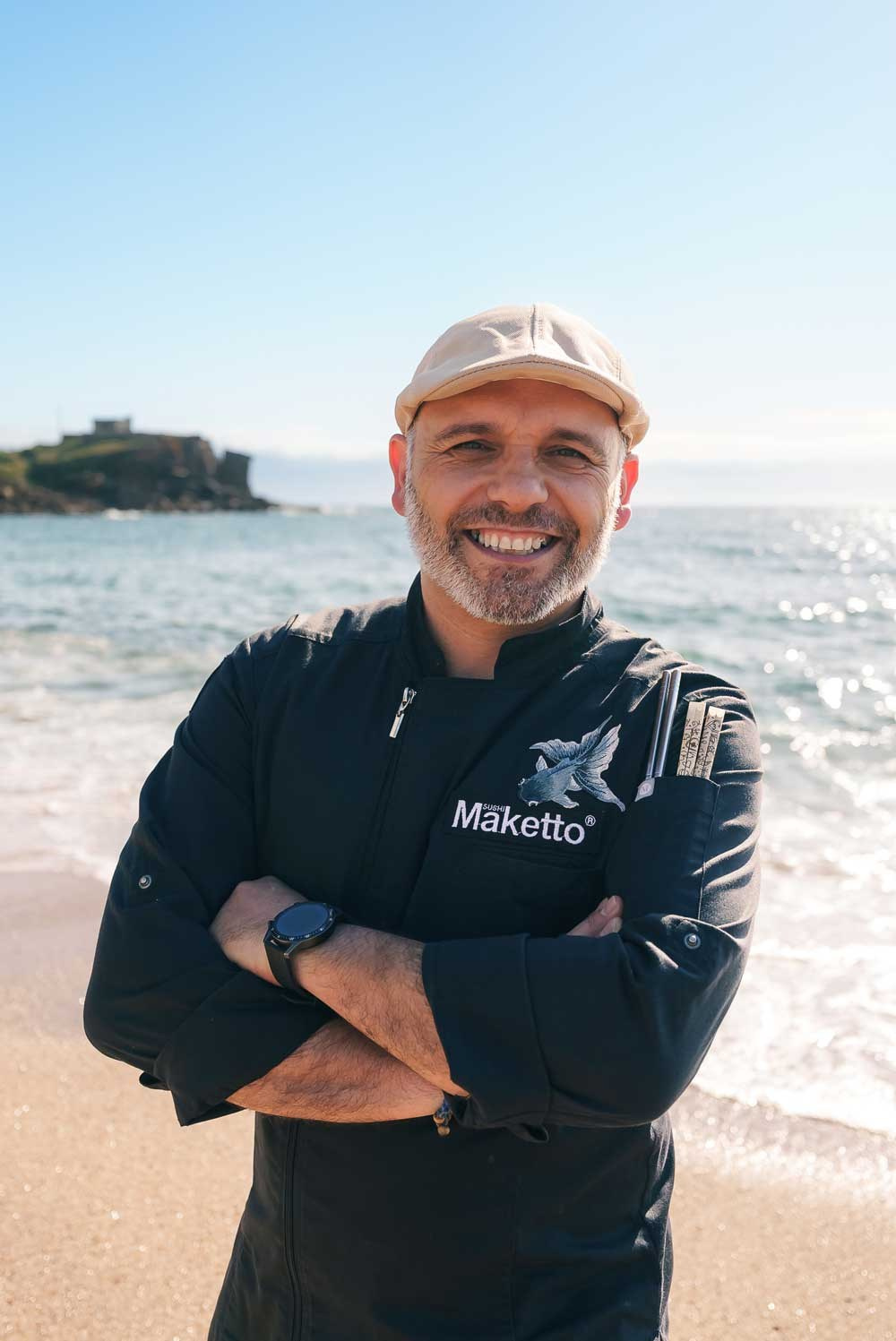 Fotografia de retrato em exterior de um homem a sorrir de braços cruzados na praia com o mar ao fundo, produzida pela Khave Media