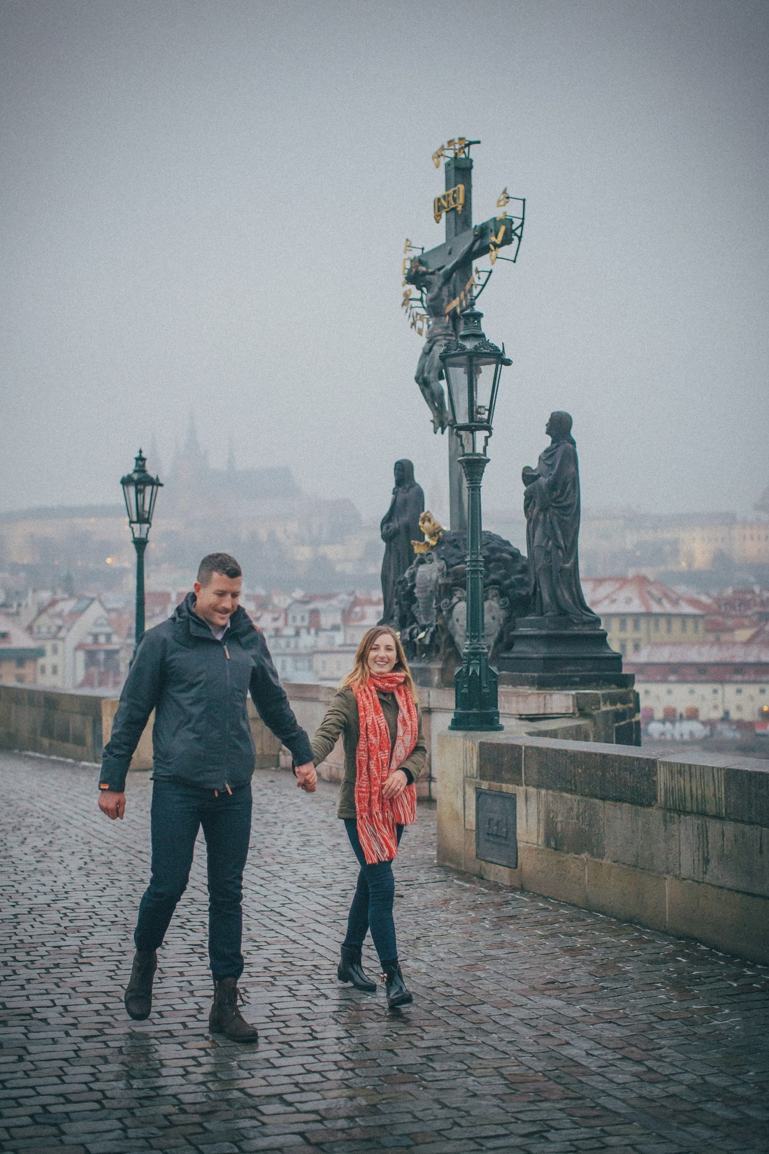 Woman wearing red scarf smiling walking hand-in-hand with partner on Charles Bridge winter morning.