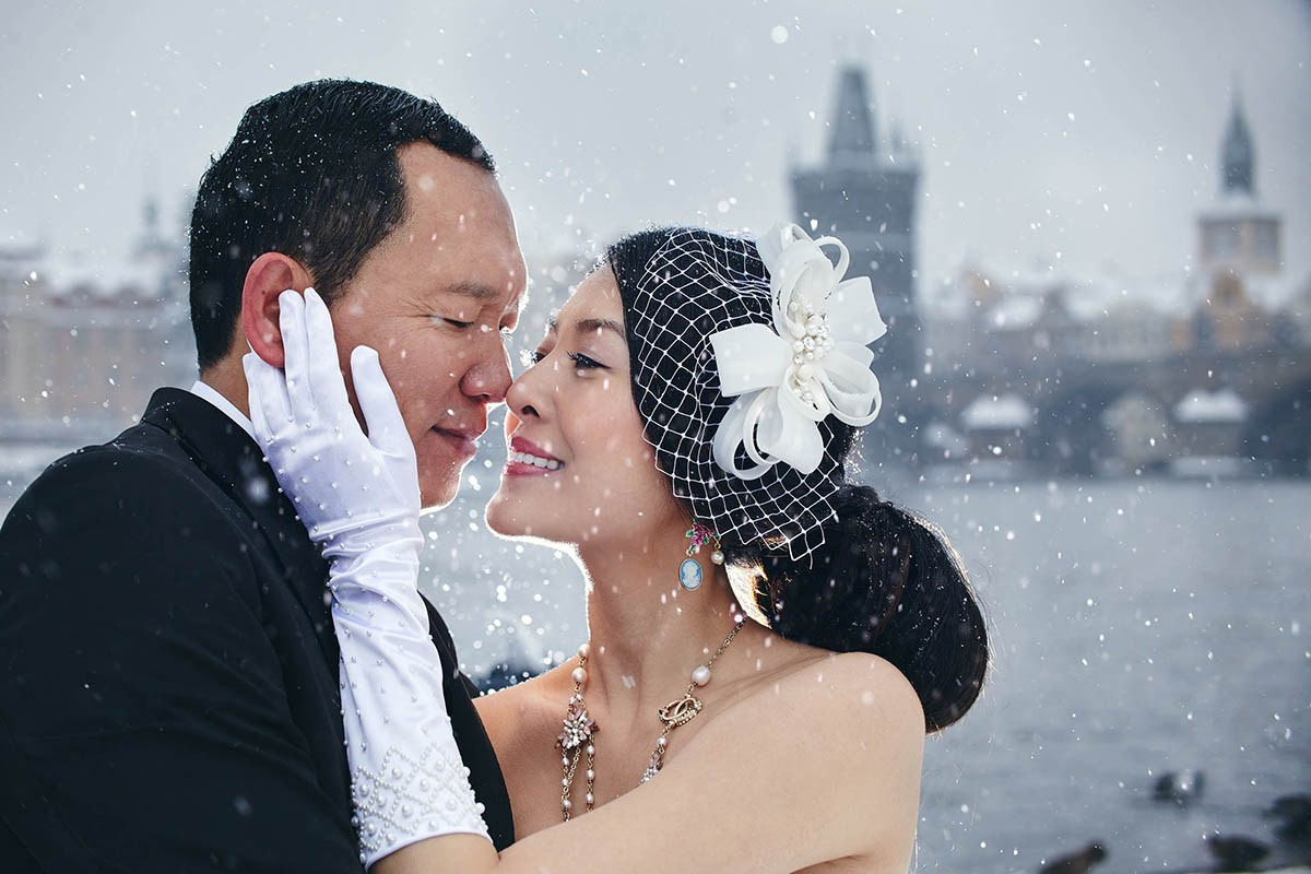 Cy and Helen embrace as the snow falls lightly around them at the riverside in Prague