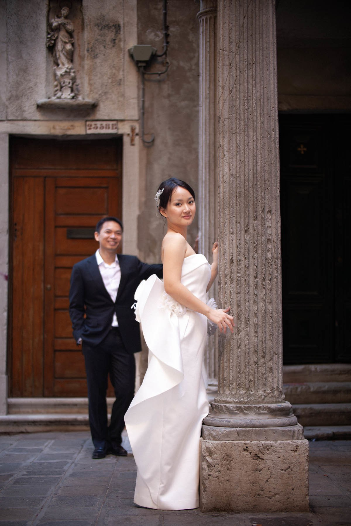 Thai bride showcasing designer dress with partner smiling behind in historic Venice quarter.