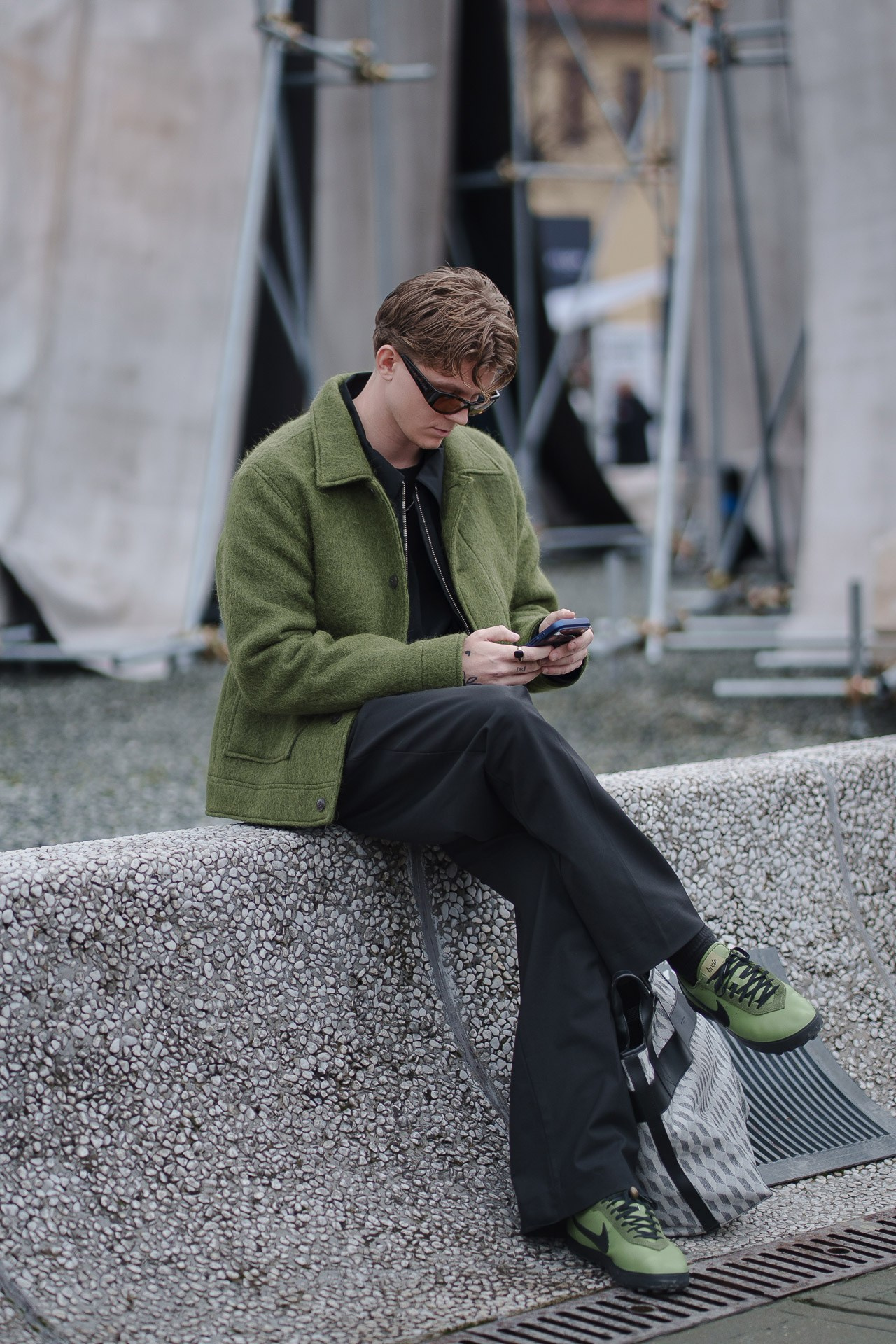 Young man sitting on concrete wall checking phone at Pitti Uomo Florence