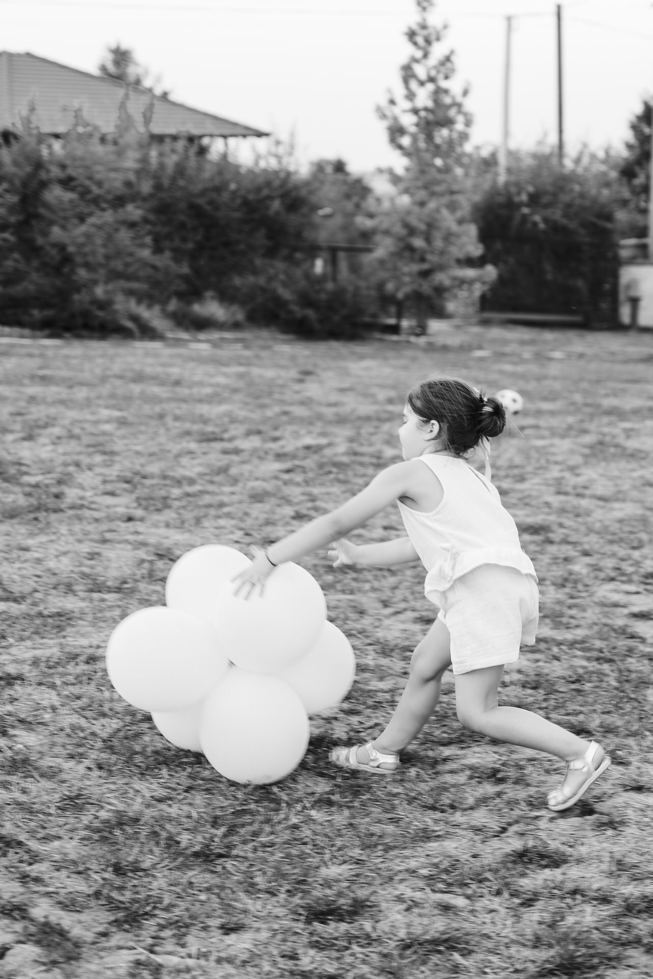 Crno-bela fotografija devojčice koja trči za grupom uvezanih balona na travnjaku.
