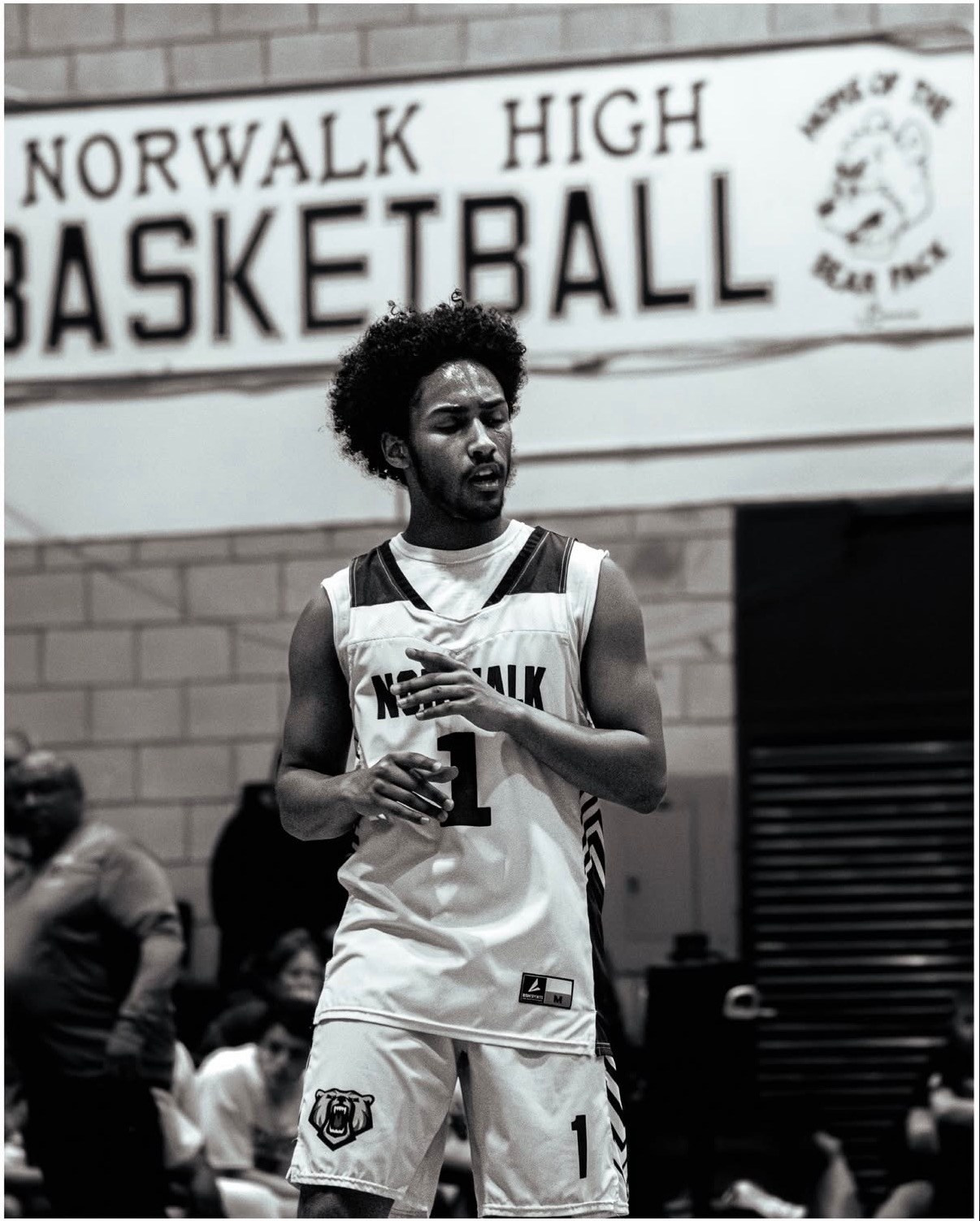 Black and white portrait of a high school basketball player standing in front of a gym basketball sign. Celebrating after scoring a 3-pointer. 