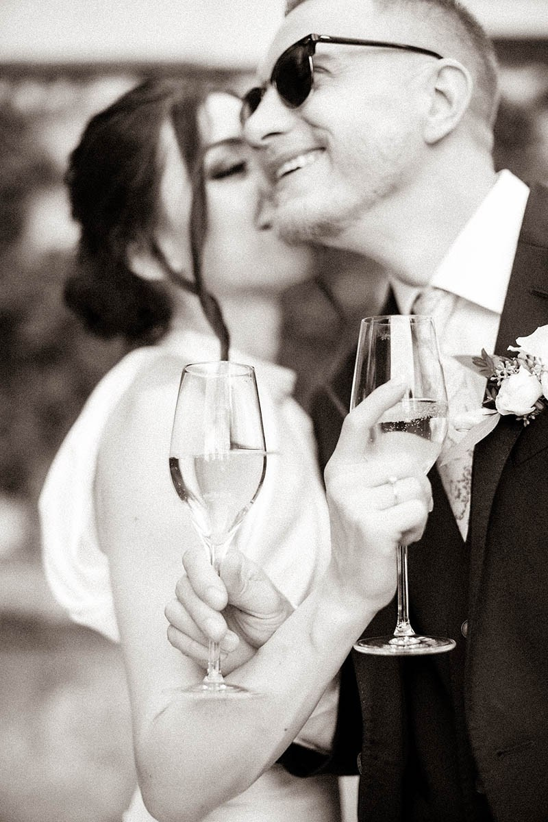 Bride smiling as groom steals a kiss while she holds a champagne glass