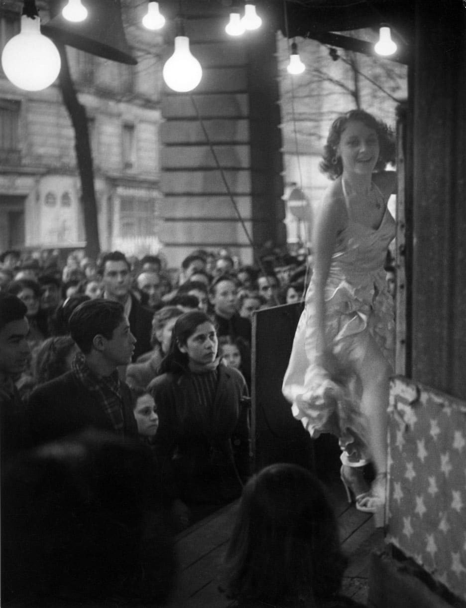 Фотография Брассая «Prostitute Undressing» (1934–1935): женщина частично раздевается в комнате с мягким освещением, передавая интимный момент из повседневной жизни.