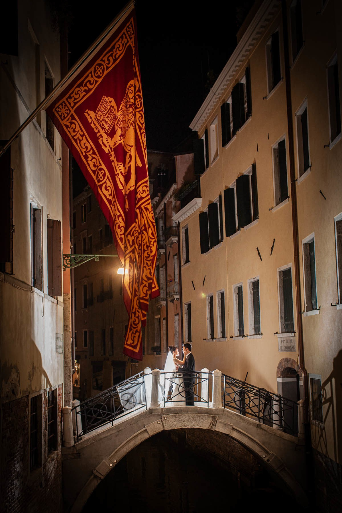 Bride and groom embracing under Banner of Saint Mark along Venice canal at night.