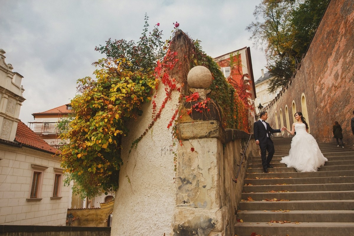A Singaporean bride walks hand-in-hand with her groom down Prague Castle steps towards the Charles Bridge as the Autumn colors guide the way