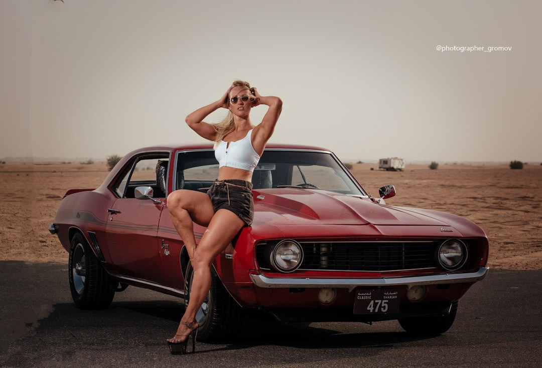 A beautiful woman sits on the bonnet of a red car against the backdrop of Dubai’s sandy roads