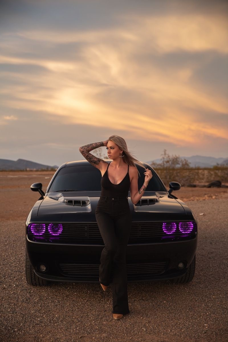 A beautiful girl is sitting on the hood of a black expensive beautiful Dodge. Against the backdrop of a beautiful Dubai sunset