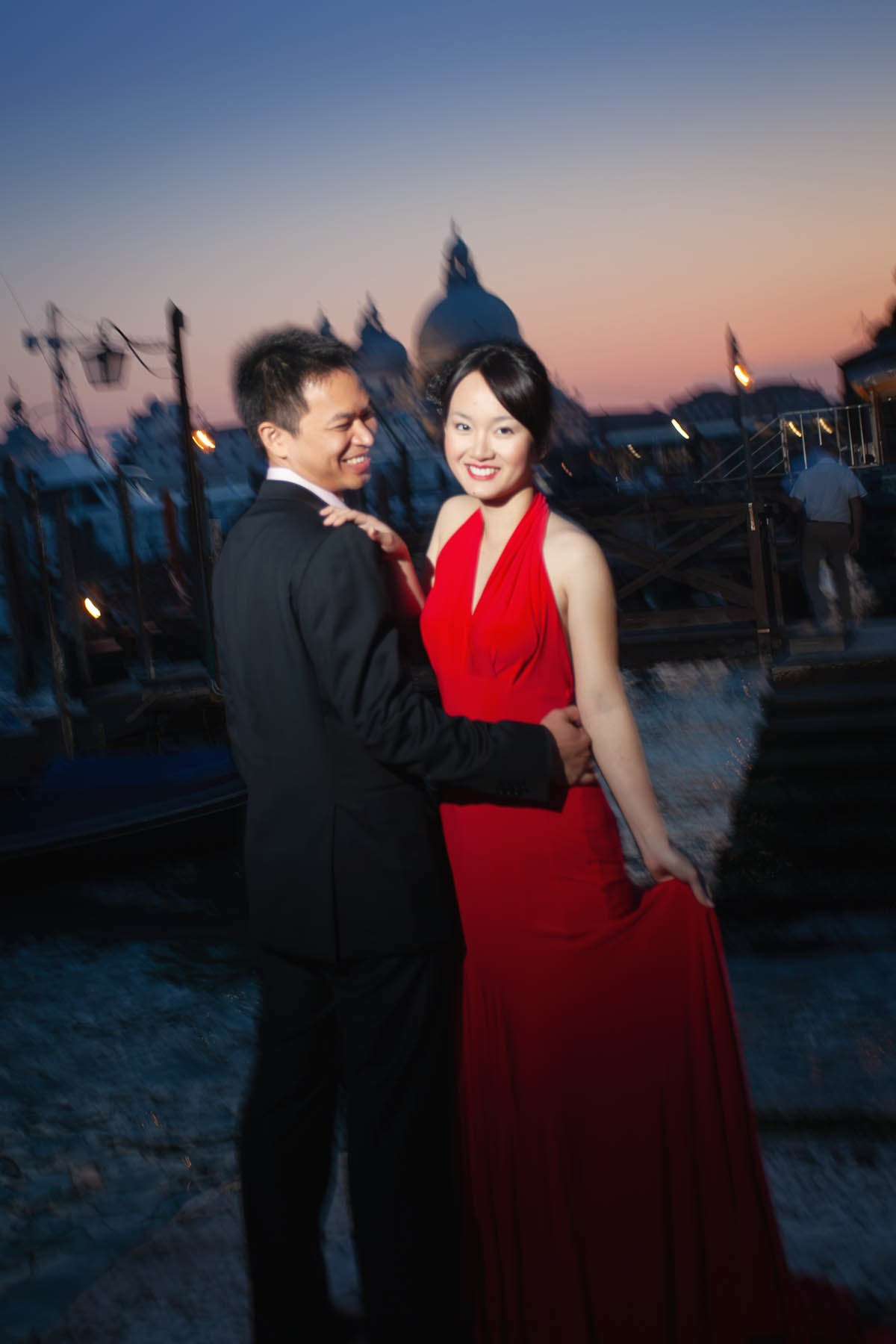 Beautiful Thai bride in skin-tight red dress smiling while held near gondola at sunset Venice.