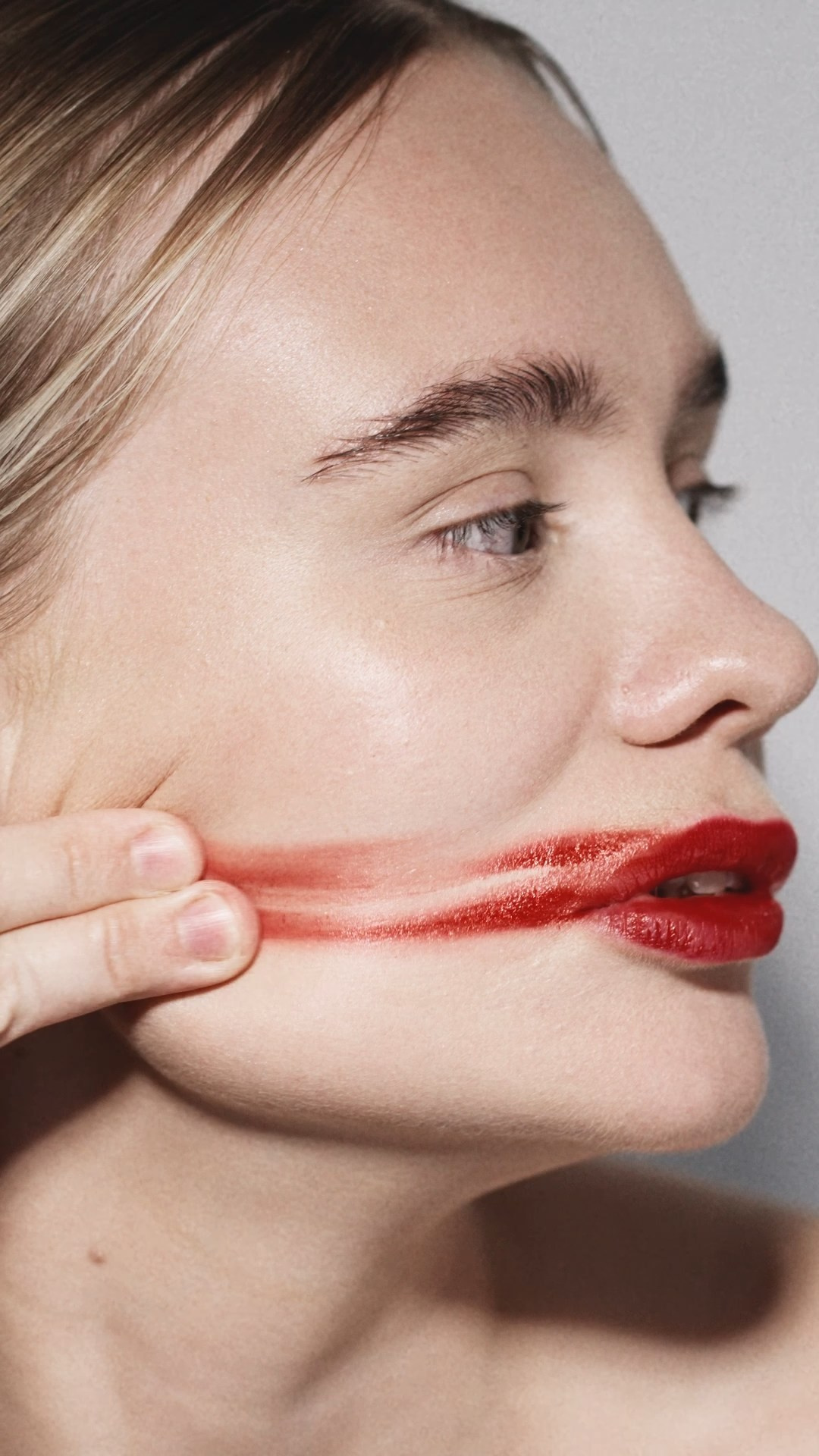  A close-up side view of a woman with red lipstick smeared across her face as she pulls her cheek, creating a striking and unconventional visual.