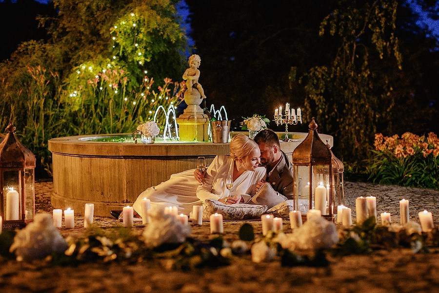 Elegant couple lying on carpets, surrounded by candles and lanterns, toasting with champagne beside a quaint fountain.