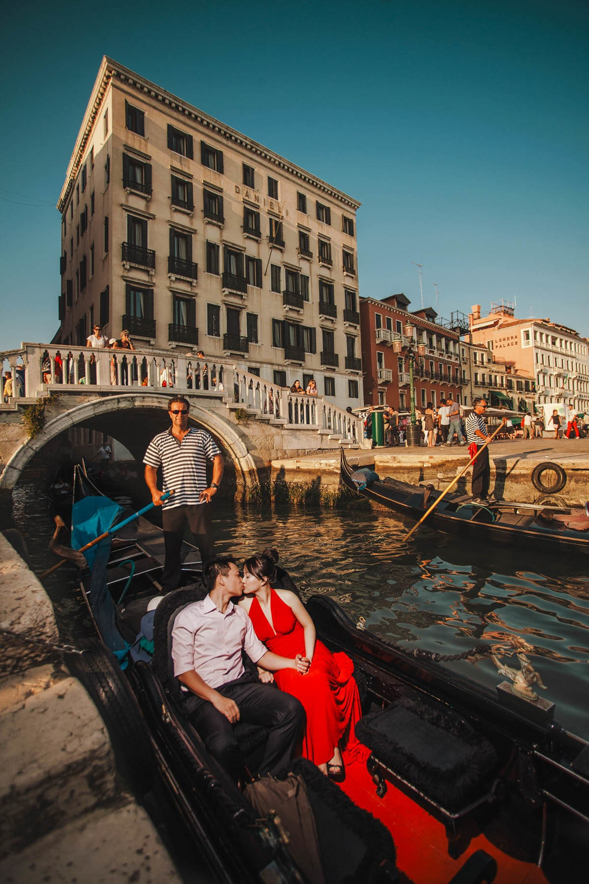 Woman in stylish red dress kissed by partner awaiting gondola near Hotel Danieli Venice.