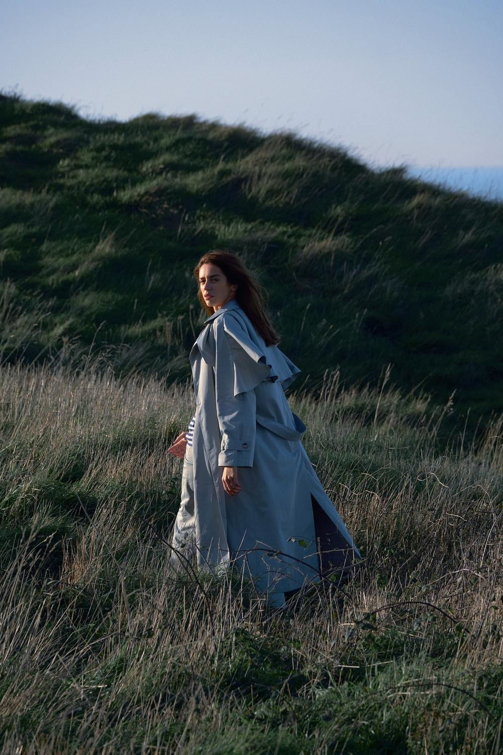 Outdoor editorial campaign with a light blue trench coat standing on a grassy hillside