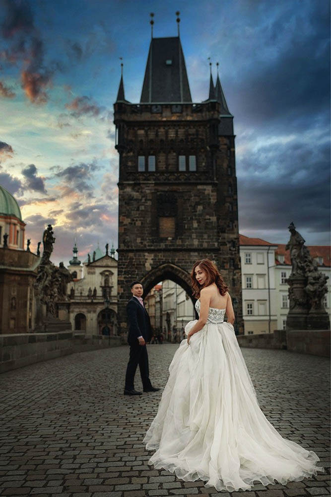 A woman in gown stands atop the Charles Bridge as her partner looks on shortly after sunrise.